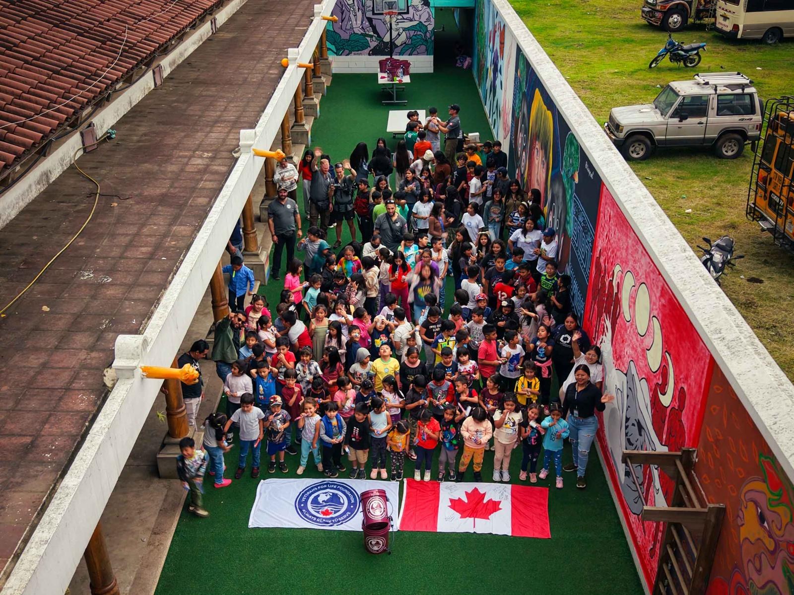 Vue aérienne d’un grand groupe d’enfants dans une cour, avec le drapeau de V4LF, le drapeau du Canada et un seau de filtration